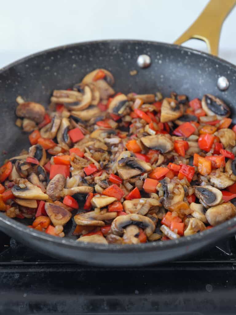 Sliced mushrooms, diced red bell peppers, and onions are being sautéed in a black frying pan with a gold handle on a stovetop.