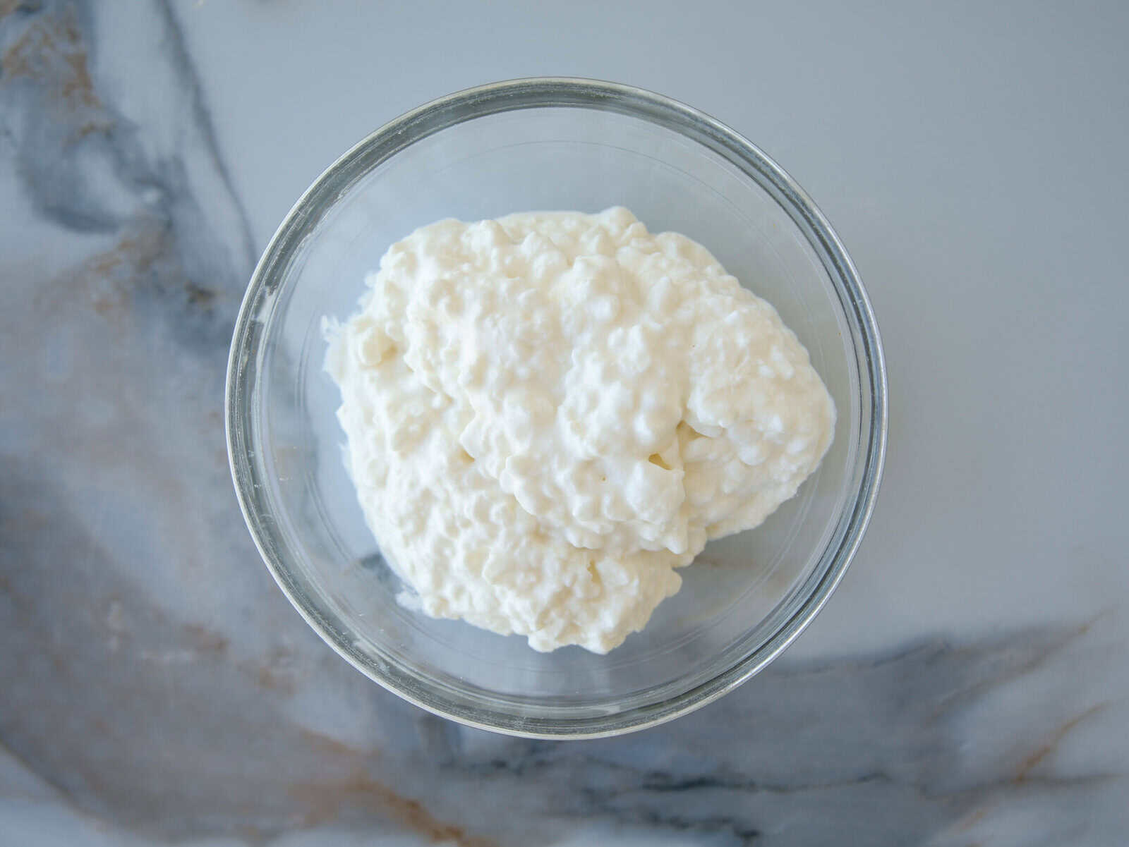 A glass bowl filled with white cottage cheese sits on a marble surface.