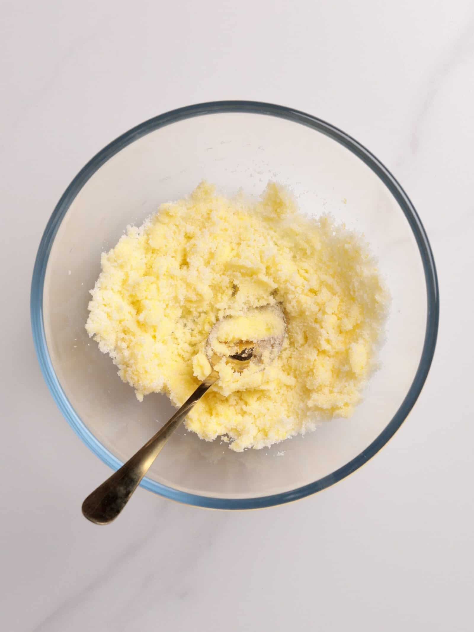 A glass bowl containing a creamy, pale yellow mixture with a metal spoon inside, sitting on a light-colored surface.