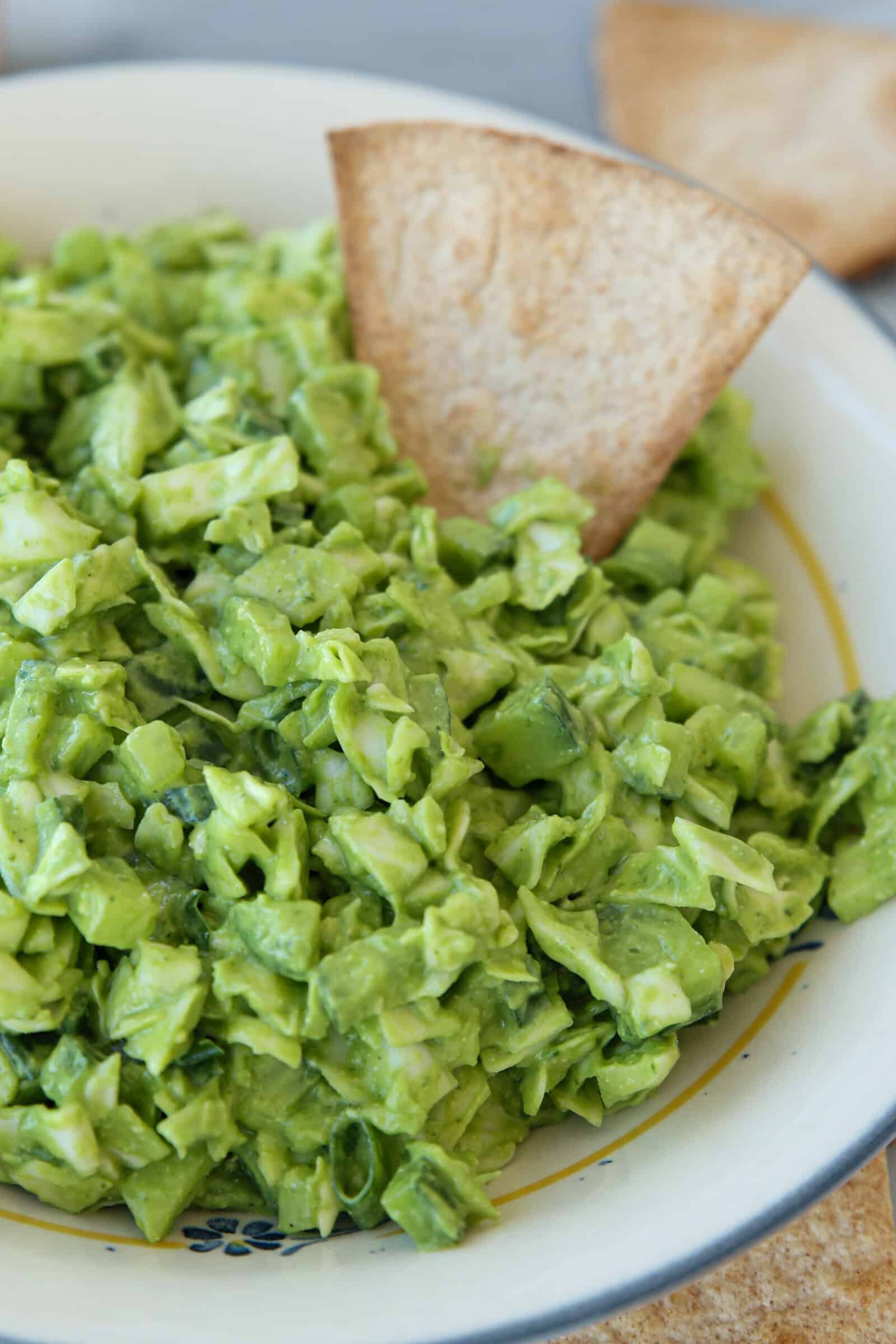 A bowl filled with chunky green goddess salad with green goddess dressing, with pieces of diced avocado and herbs, garnished with a single triangular tortilla chip placed upright in the dip.