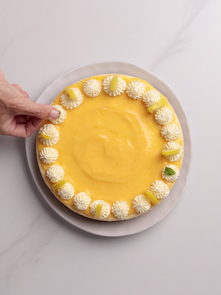 A hand places a garnish on a round lemon cheesecake topped with piped whipped cream, lemon slices, and mint leaves, displayed on a white plate against a light background.