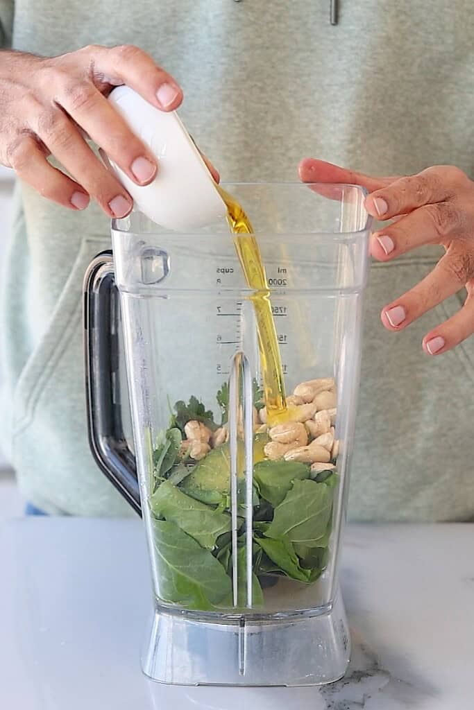 A person pours olive oil from a small white cup into a blender filled with green leaves and cashew nuts.
