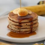 A stack of four cottage cheese and banana pancakes topped with banana slices, with syrup being poured over them, sits on a white plate with a blurred banana in the background.
