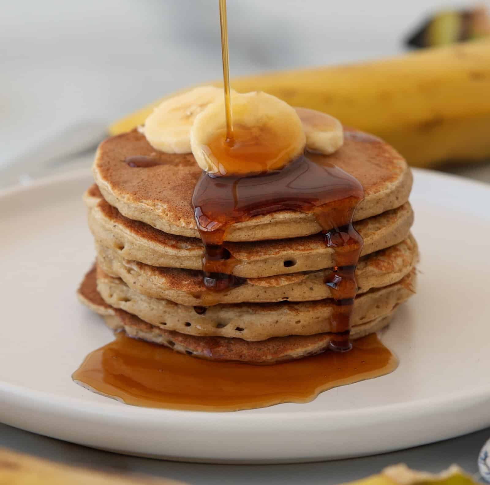 A stack of four cottage cheese and banana pancakes topped with banana slices, with syrup being poured over them, sits on a white plate with a blurred banana in the background.