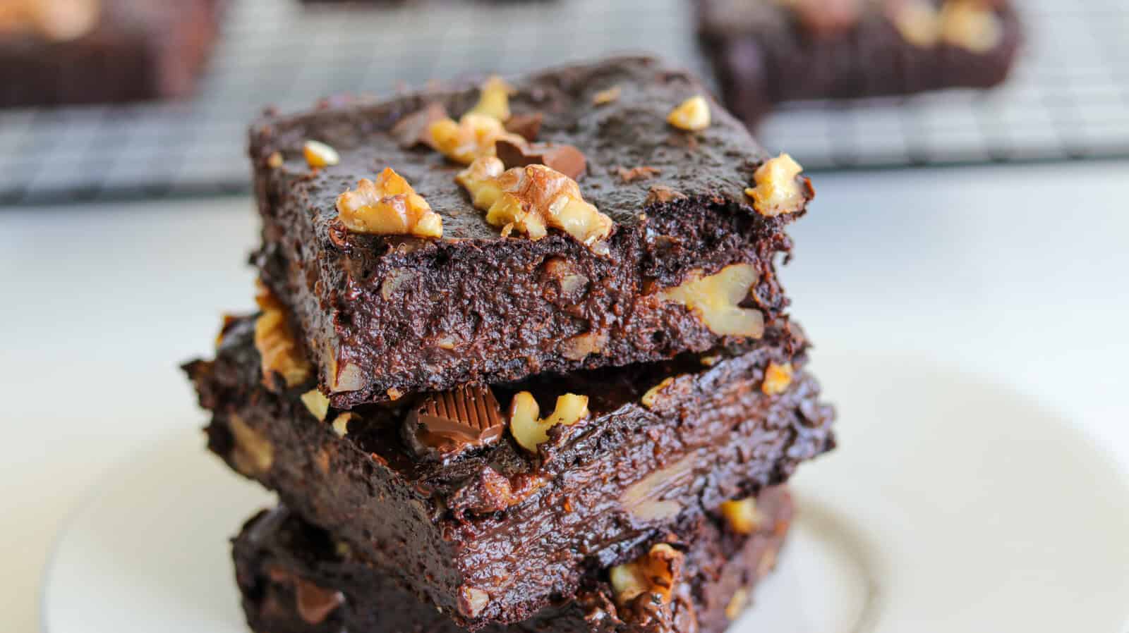 A close-up of three thick, fudgy brownies stacked on a white plate. The brownies are studded with chopped walnuts and chocolate chunks, and a cooling rack with more brownies is blurred in the background.