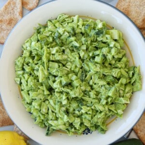 A bowl filled with a creamy green chopped salad, surrounded by pita chips and a partial view of a lemon and avocado on a white surface.
