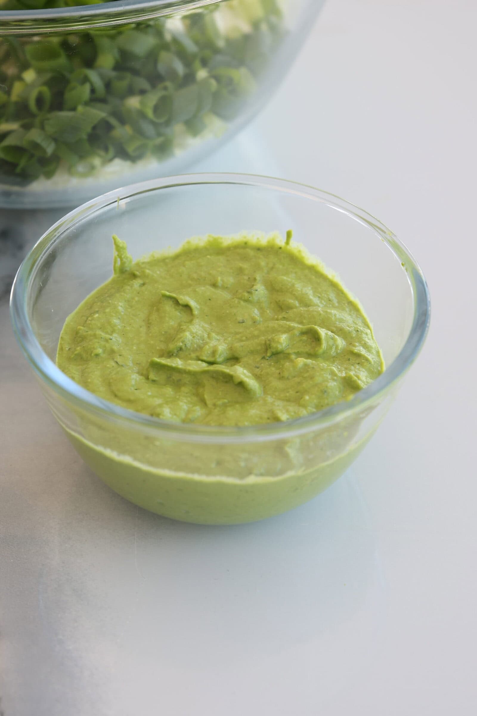 A clear glass bowl filled with creamy green goddess dressing sits on a white surface. In the background, another bowl contains chopped green onions.