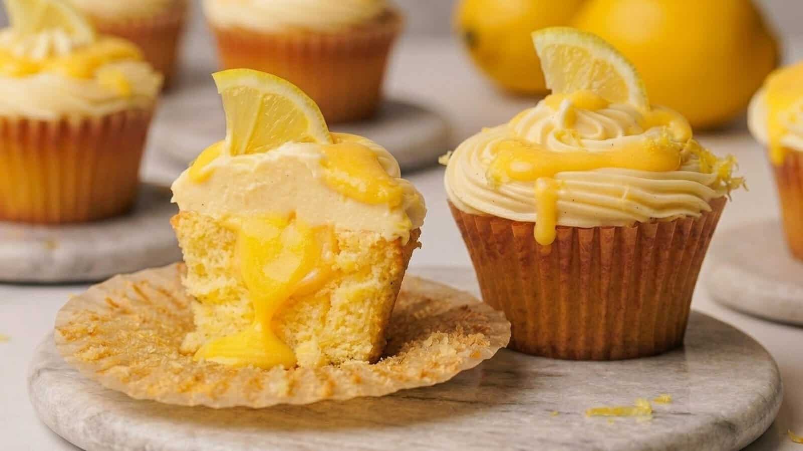 Two lemon cupcakes with creamy frosting and lemon drizzle, one cut in half to show a gooey center. Each cupcake is topped with a slice of lemon. Whole lemons and more cupcakes appear in the blurred background.