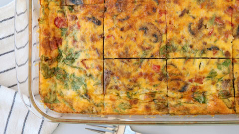 A baked vegetable cottage cheese egg casserole is sliced into squares in a glass dish, showing pieces of spinach, mushrooms, and tomatoes. A striped kitchen towel and a metal serving utensil are nearby.