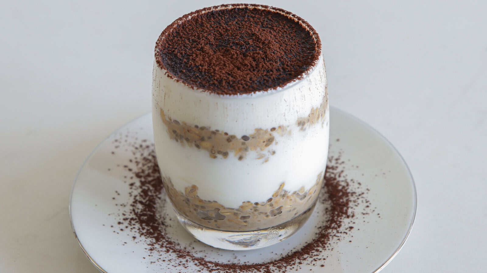 A glass of layered tiramisu overnight oats with creamy white and coffee-soaked layers, topped with cocoa powder, sits on a white plate dusted with cocoa.