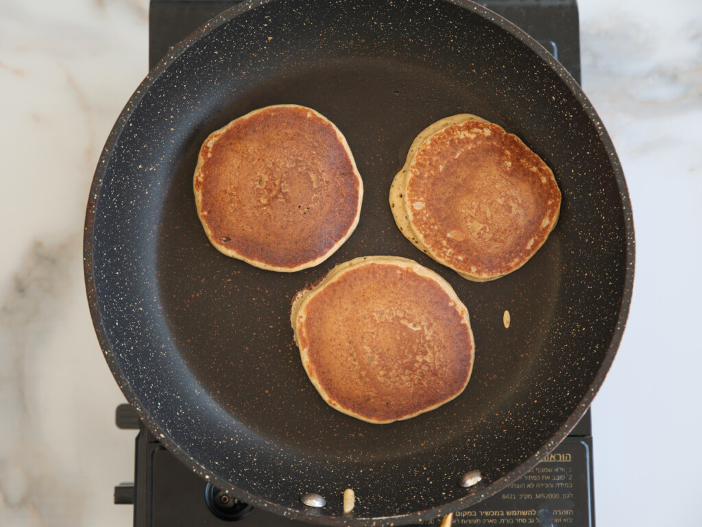 Three golden-brown pancakes are cooking in a black nonstick frying pan on a stovetop, viewed from above. The pancakes are evenly spaced with slight browning on their surfaces.