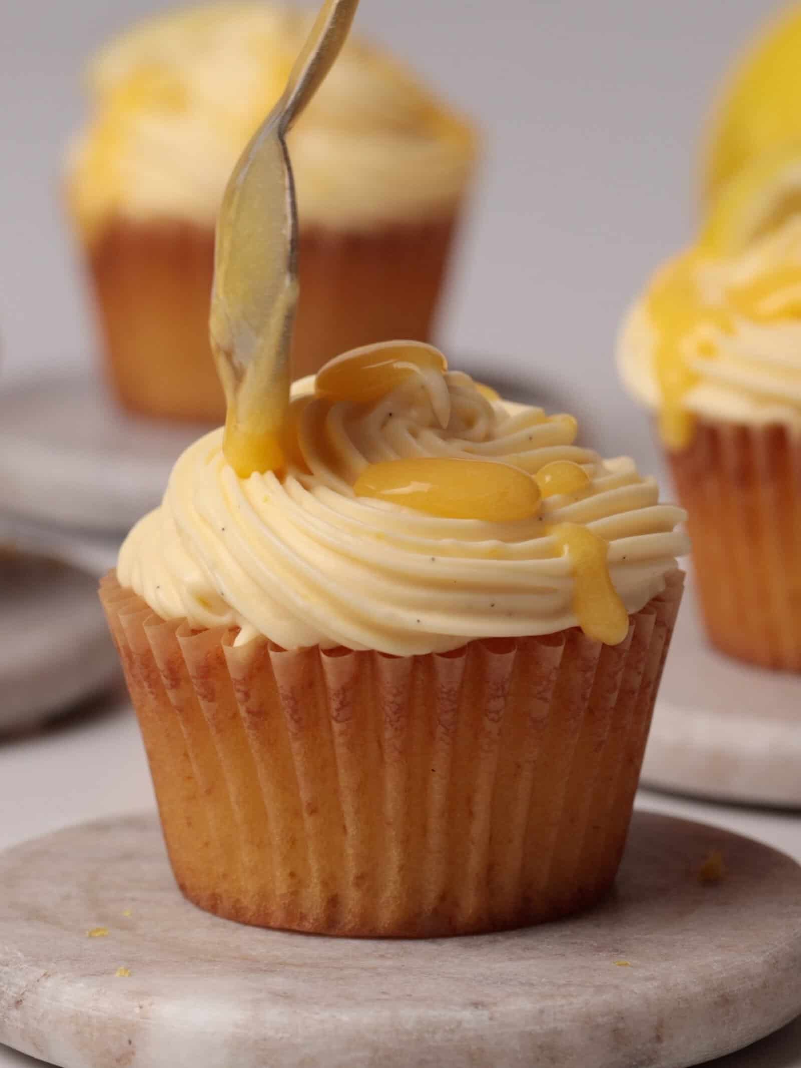 A lemon cupcake with swirled lemon frosting is being topped with lemon curd from a spoon, sitting on a small round marble plate. Another frosted cupcake is visible in the background.