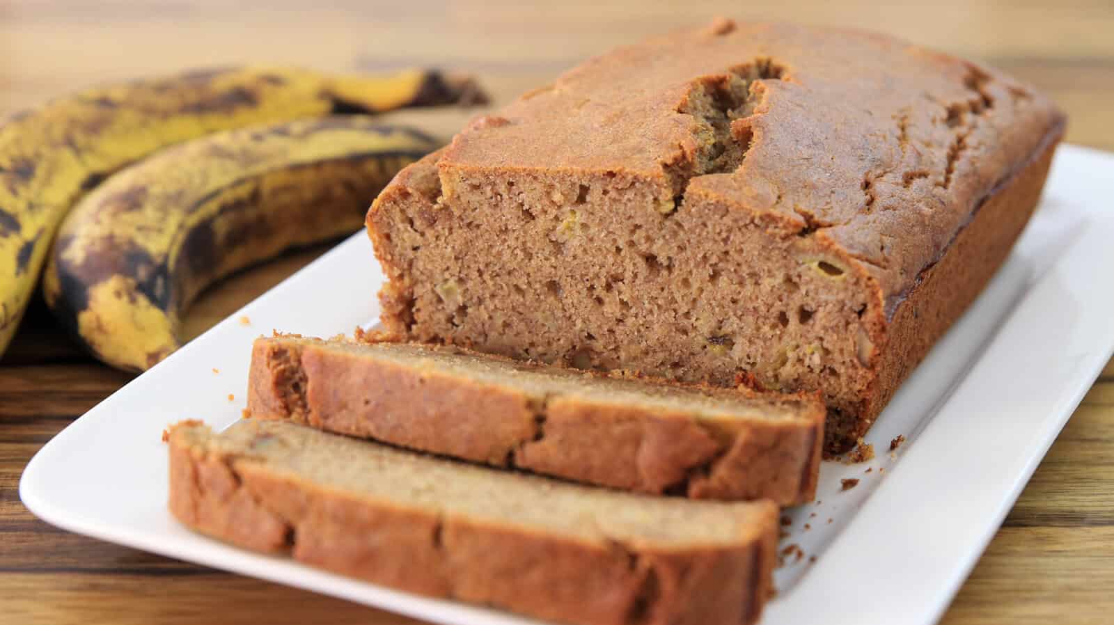 A loaf of healthy banana bread with two slices cut sits on a white plate. Two ripe bananas are in the background on a wooden surface.