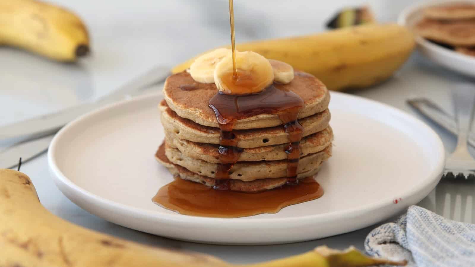 A stack of cottage cheese banana pancakes topped with banana slices is being drizzled with syrup on a white plate, surrounded by whole bananas.