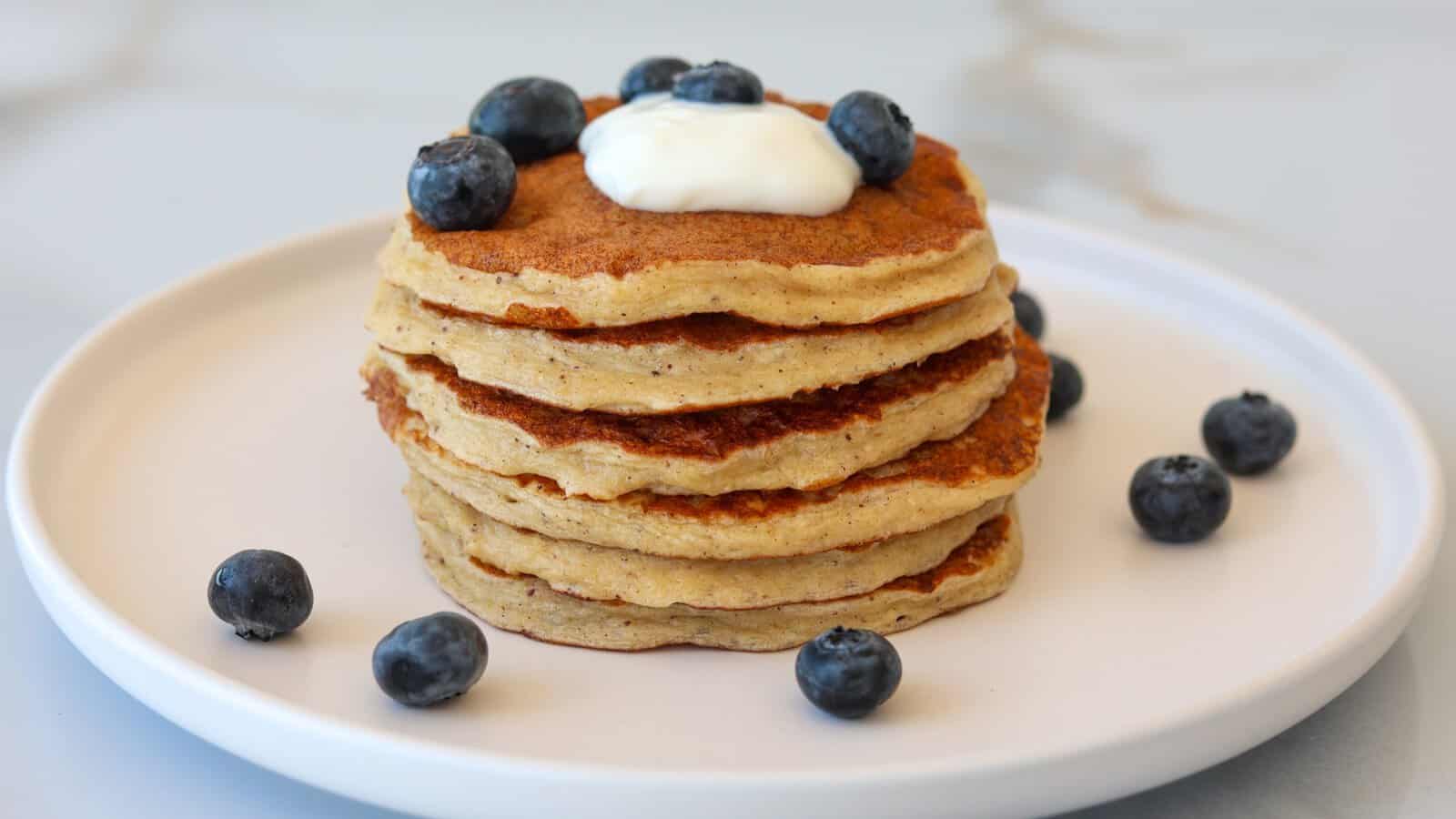 A stack of four pancakes topped with a dollop of yogurt or cream and fresh blueberries, served on a white plate with extra blueberries scattered around.