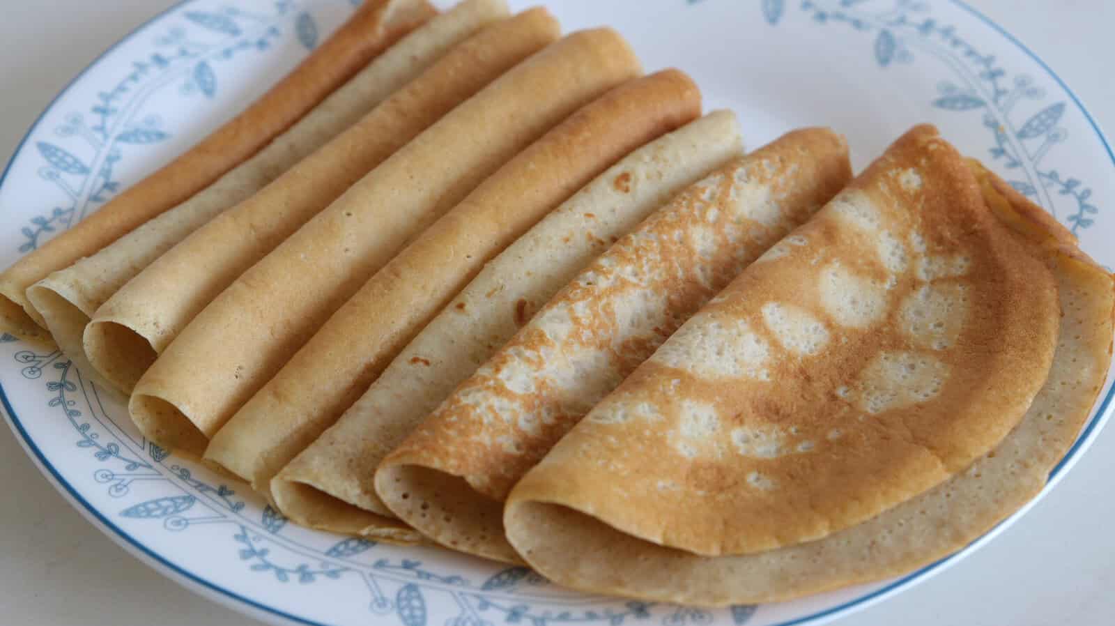 A plate with several thin, golden-brown oatmeal crepes neatly folded and arranged side by side, displaying their soft and slightly crispy texture. The plate has a blue floral pattern around the rim.