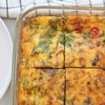A glass baking dish with a baked egg casserole with cottage cheese, topped with melted cheese, spinach, and visible vegetables, cut into four square pieces. A white plate and a checkered cloth are partially visible nearby.