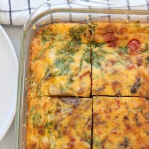 A glass baking dish with a baked egg casserole with cottage cheese, topped with melted cheese, spinach, and visible vegetables, cut into four square pieces. A white plate and a checkered cloth are partially visible nearby.