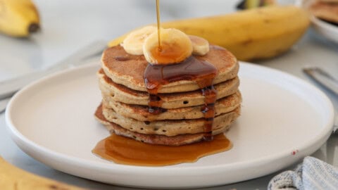 A stack of banana cottage cheese pancakes topped with banana slices and syrup being poured over them, set on a white plate with whole bananas and cutlery in the background.