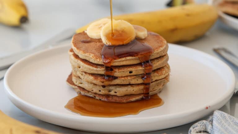 A stack of banana cottage cheese pancakes topped with banana slices and syrup being poured over them, set on a white plate with whole bananas and cutlery in the background.