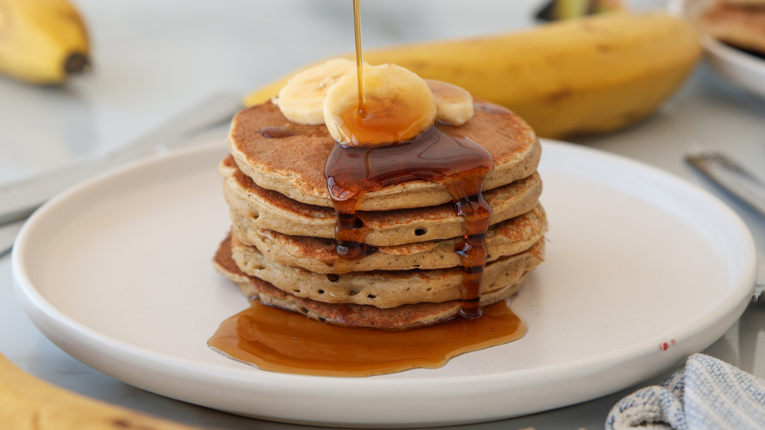 A stack of banana cottage cheese pancakes topped with banana slices and syrup being poured over them, set on a white plate with whole bananas and cutlery in the background.
