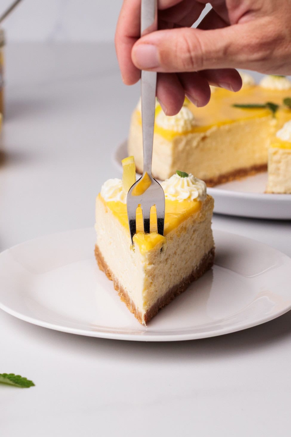 A hand holds a fork, cutting into a slice of cheesecake topped with whipped cream and yellow fruit glaze, placed on a white plate.
