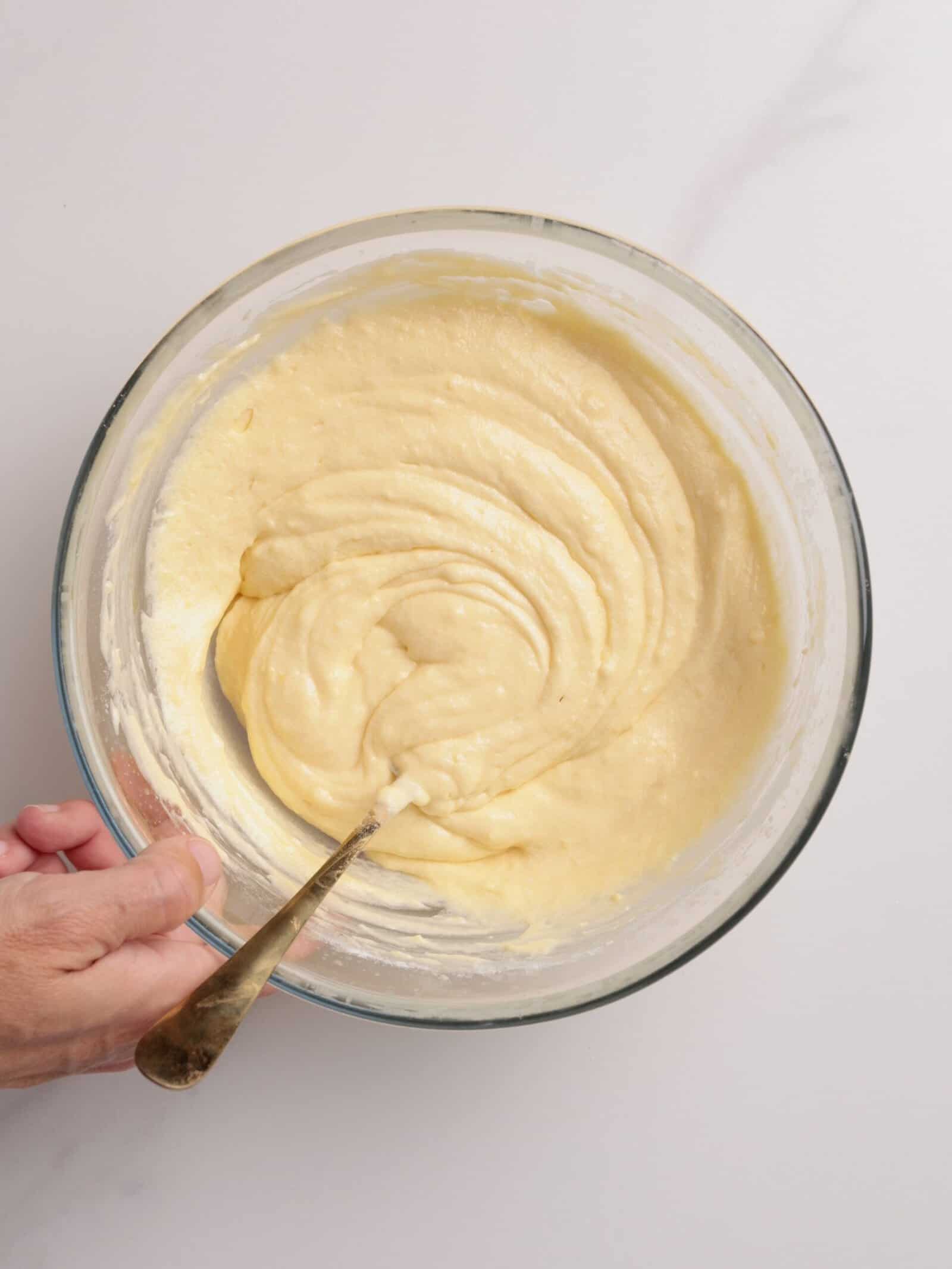 A hand holding a glass bowl filled with smooth, creamy lemon cupcake batter being mixed with a spoon, on a light-colored surface.