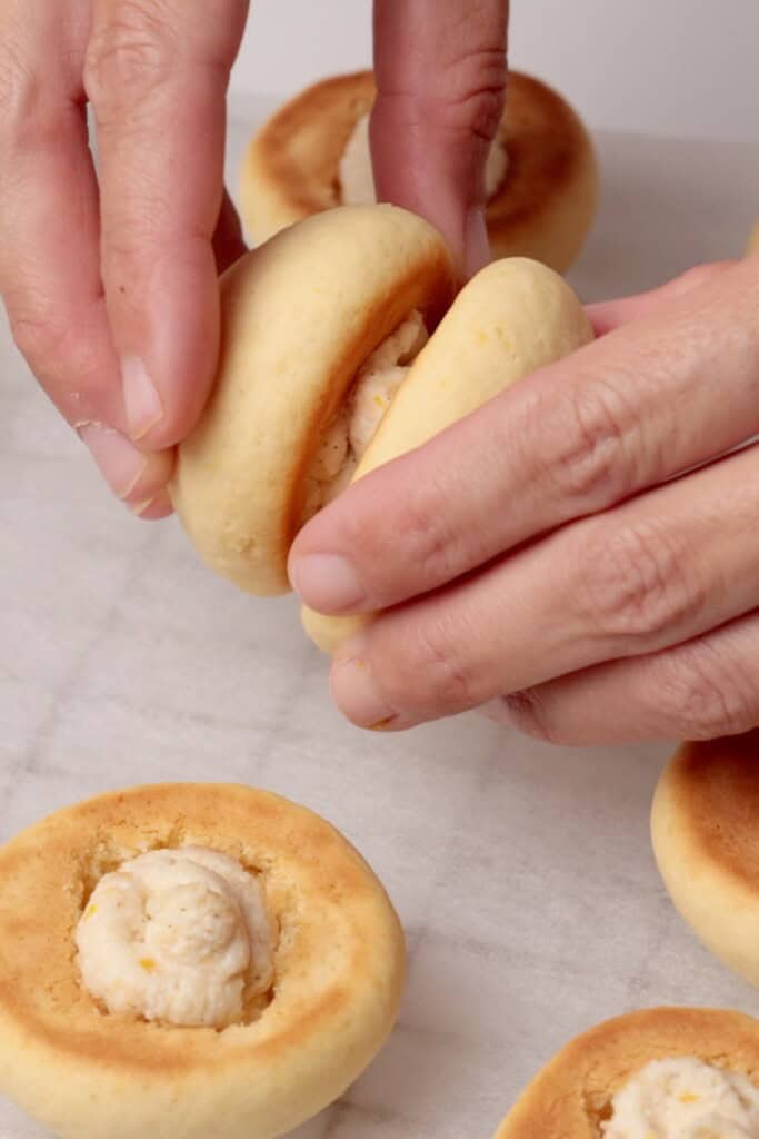 Close-up of hands assembling sandwich cookies with a creamy filling on a light surface; several cookies are visible, some stacked and some lying flat.