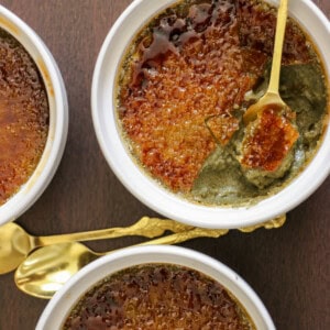 Three white ramekins of crème brûlée with caramelized sugar tops are shown. One has a spoon breaking the crust, revealing a creamy dark filling. Two gold spoons lie next to the dishes on a dark wooden surface.