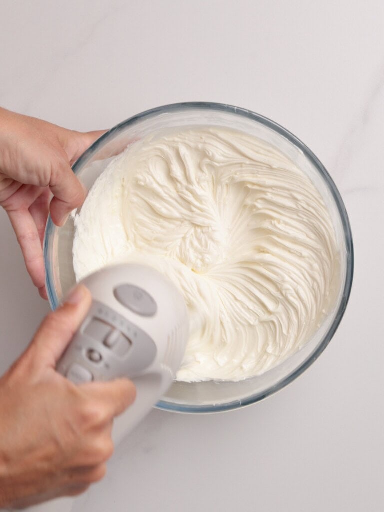 A person uses a handheld electric mixer to whip cream in a clear glass bowl on a white surface. The cream is thick and forms soft peaks.