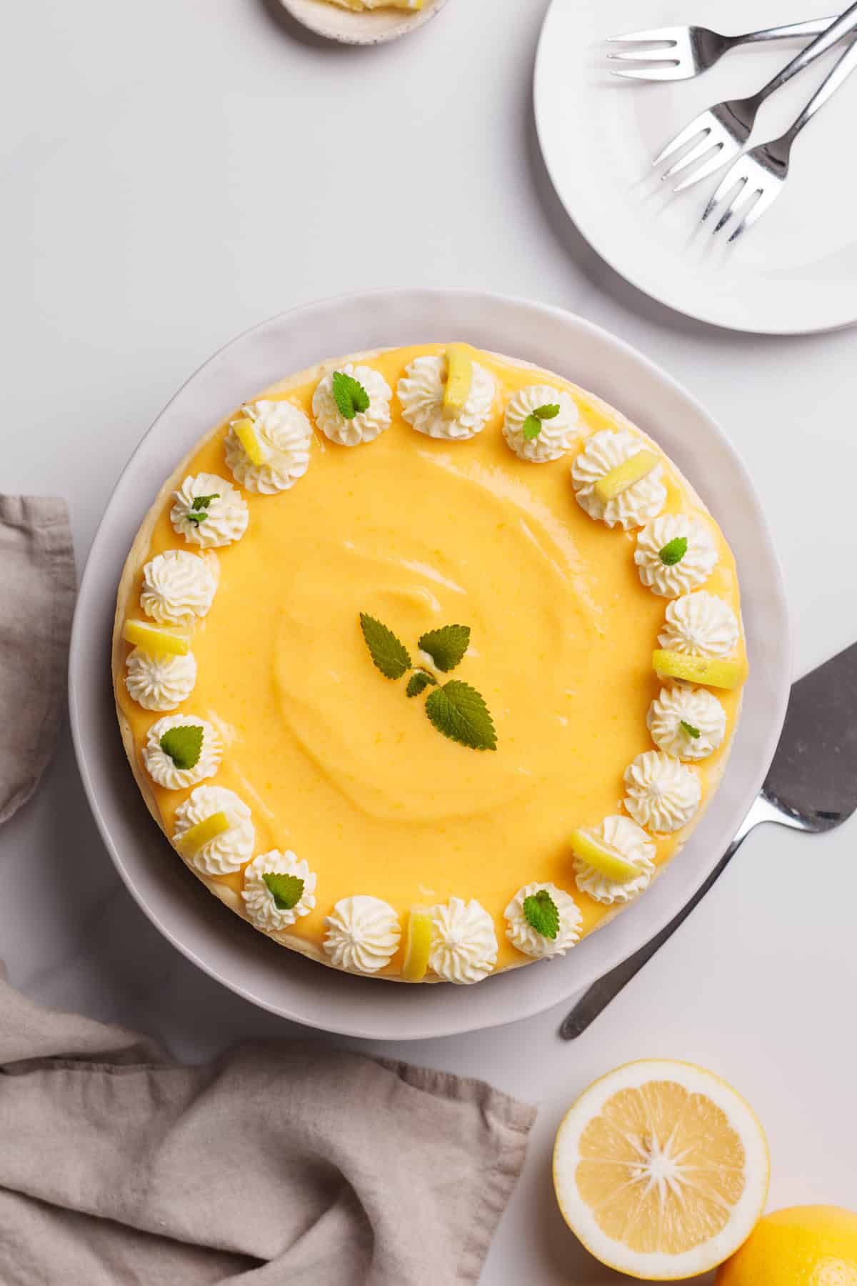 A round lemon cheesecake decorated with whipped cream, lemon slices, and mint leaves sits on a white plate. A cake server, forks, and a halved lemon are nearby on a light background.