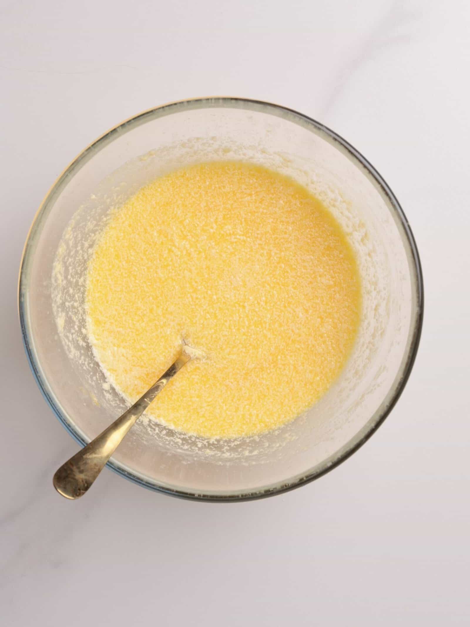 A glass bowl filled with a yellow, grainy mixture and a silver spoon resting inside, placed on a light-colored surface.