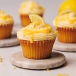 A lemon cupcake with lemon frosting, lemon zest, and a small lemon wedge on top sits on a round marble coaster. More decorated cupcakes and a whole lemon are blurred in the background.
