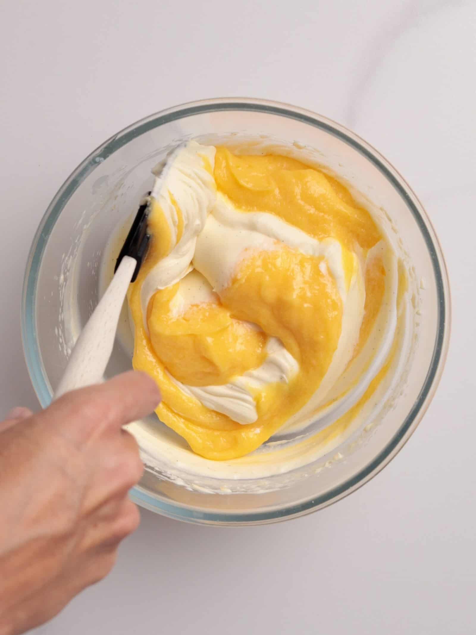 A hand uses a spatula to gently fold yellow egg mixture into a bowl of white cream, blending the ingredients together on a light surface.