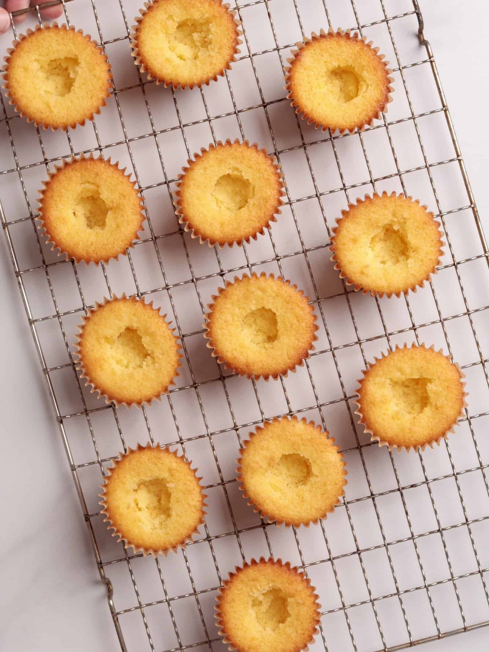 A cooling rack holds twelve yellow cupcakes, each with a hole in the center, arranged diagonally on a white surface.