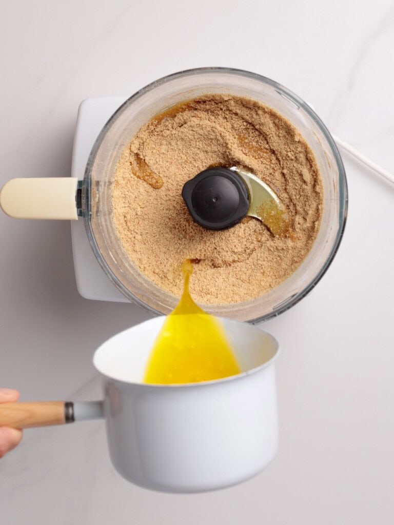 A hand pours melted butter from a white saucepan into a food processor containing crushed graham crackers, seen from above on a light countertop.