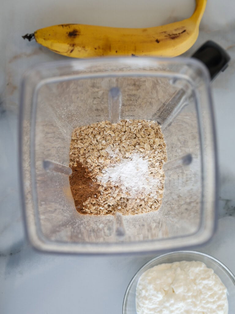 A top-down view of a blender containing oats, cinnamon, and baking powder. Above the blender is a whole banana, and below it is a bowl of cottage cheese.