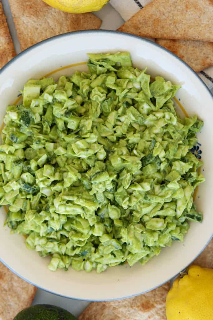 A bowl filled with creamy, chopped avocado salad sits on a white plate, surrounded by pita bread slices and a lemon. The salad appears fresh and vibrant green.