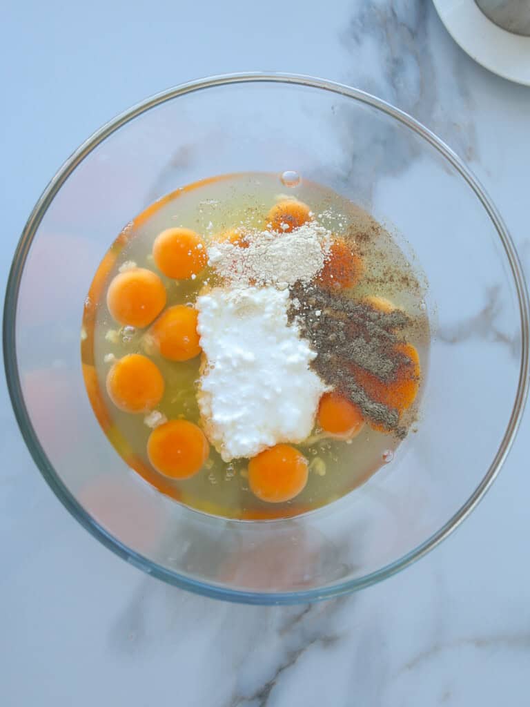 A glass bowl containing cracked eggs, garlic powder, cottage cheese, salt, black pepper, and paprika on a white marble countertop.