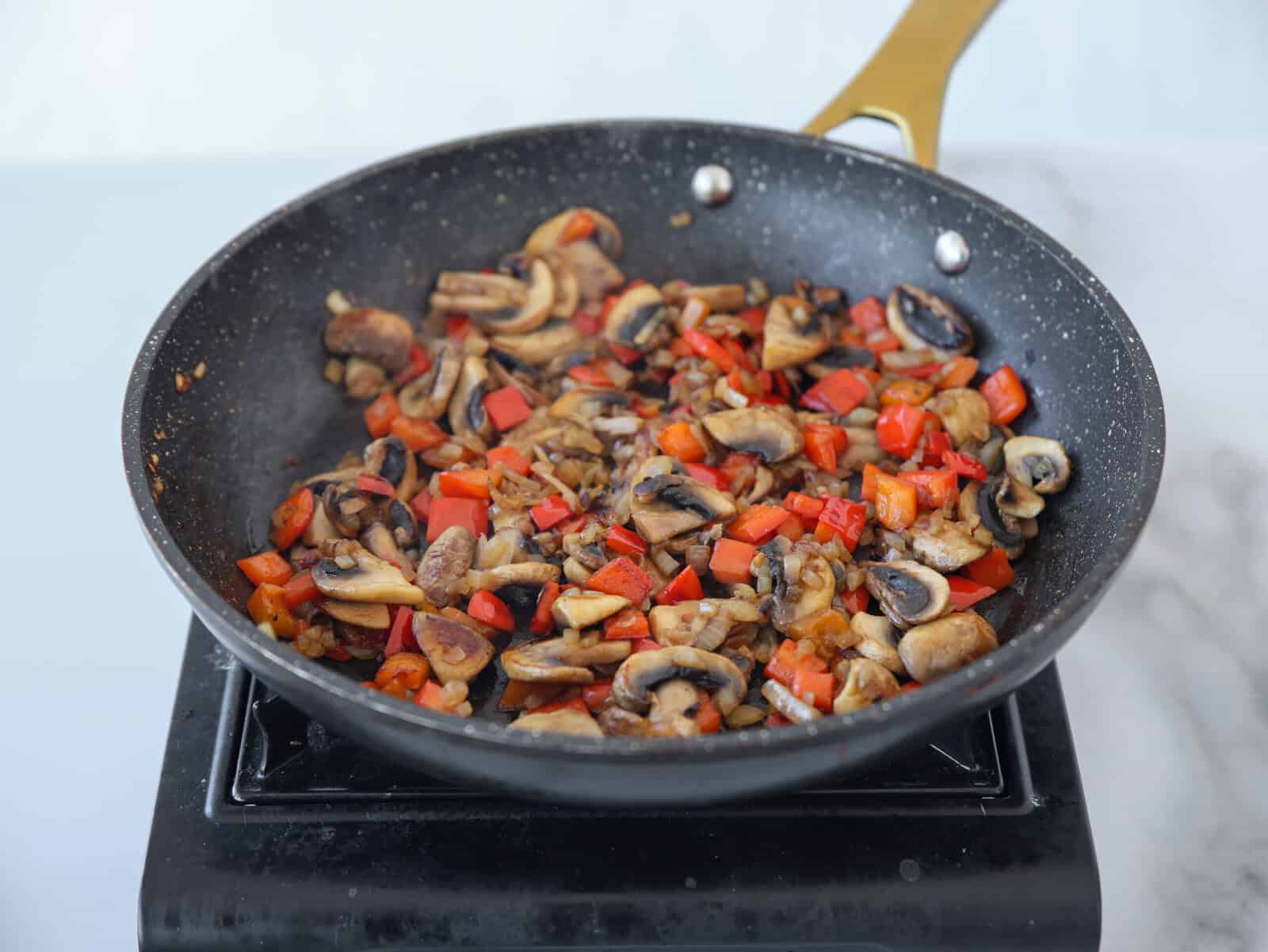 Sliced mushrooms, red bell peppers, and onions cooking in a black frying pan with a gold handle on a stovetop. Steam rises from the sautéed vegetables.