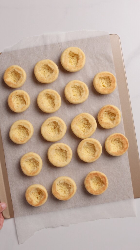 A baking tray lined with parchment paper holds 19 baked cookie cups, each with a hollow center, arranged in neat rows on a white surface.
