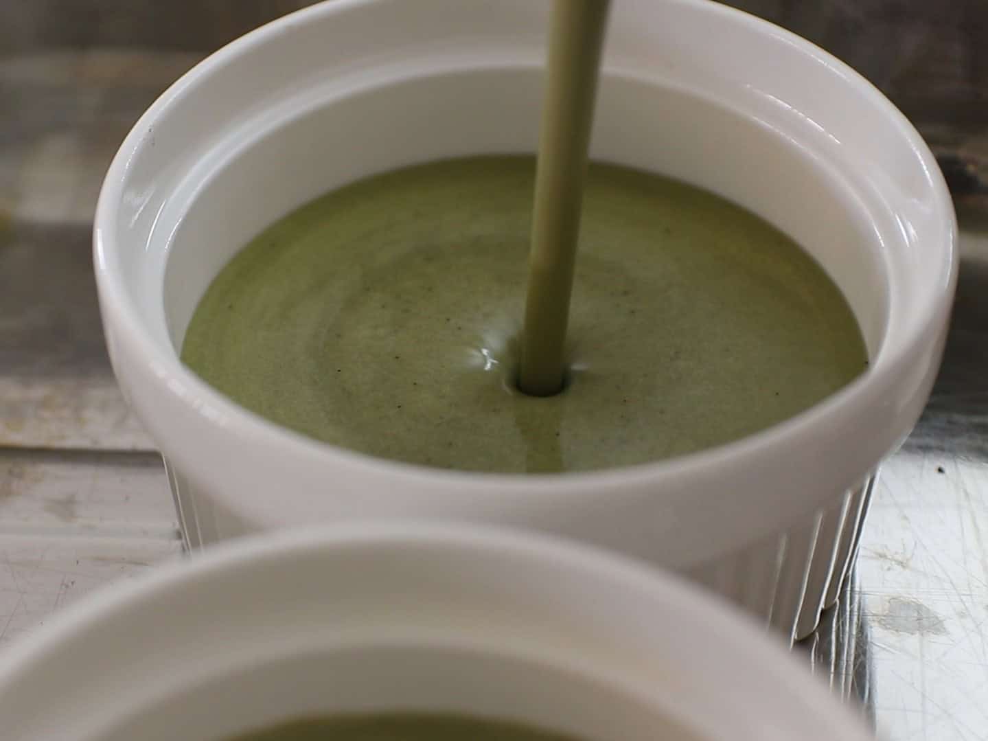 Green pistachio custard is being poured into a white ramekin, filling it nearly to the top. Another similar ramekin is partially visible in the foreground. The surface is metallic and reflective.