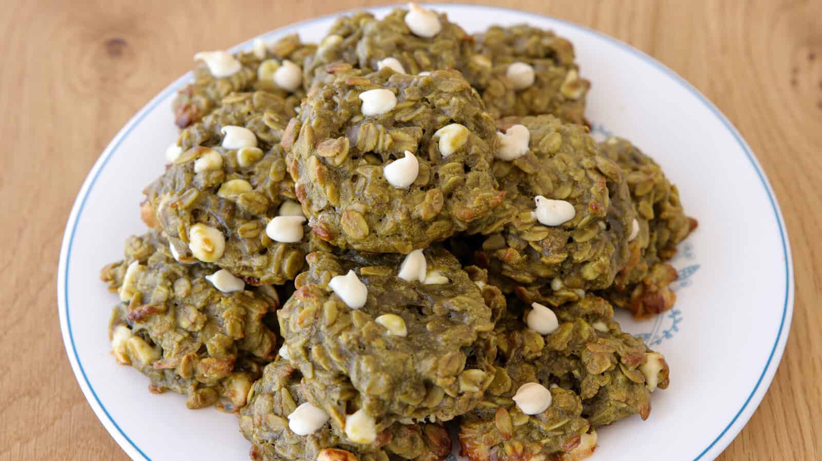 A plate of green oatmeal cookies with white chocolate chips, arranged in a pile on a white plate with a blue rim, set on a wooden surface.