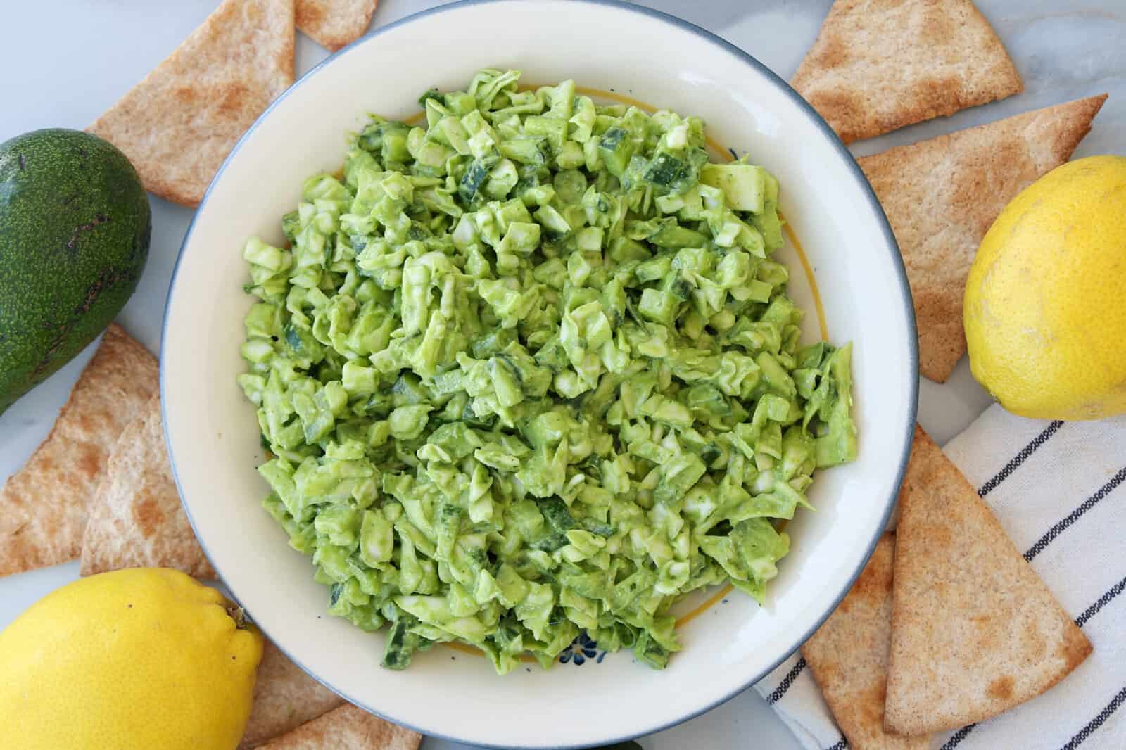 A bowl of chopped green goddess salad, with avocado, and creamy green goddess dressing is surrounded by pita chips, a lemon, an avocado, and a striped kitchen towel on a marble surface.