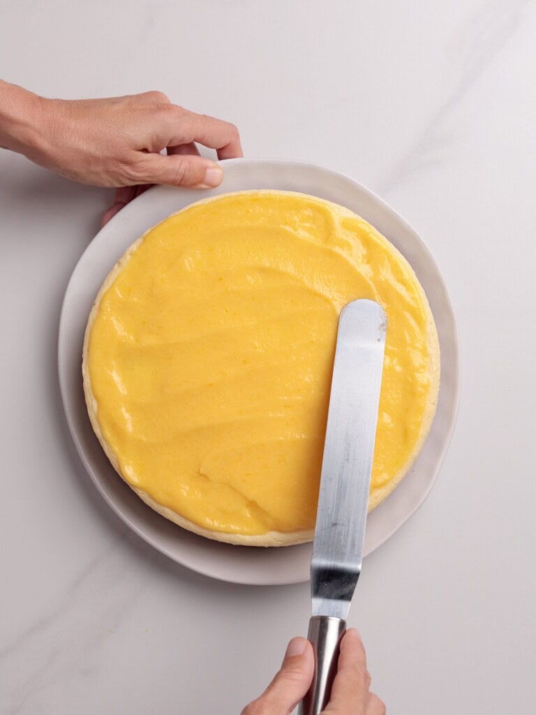 A person spreads yellow frosting smoothly over a round cake with an icing spatula, while holding the white plate steady on a light-colored surface.