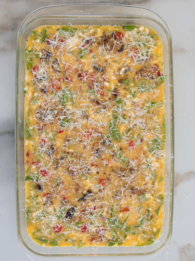 A glass baking dish filled with an unbaked egg mixture containing chopped vegetables, visible greens, red peppers, and topped with shredded cheese, ready to be cooked.