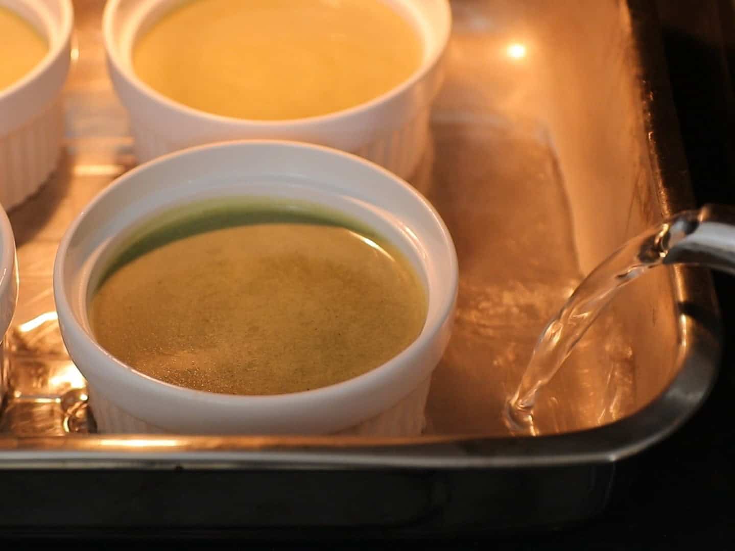 A close-up of white ramekins filled with green custard mixture in a metal baking tray, with hot water being poured into the tray for a water bath.