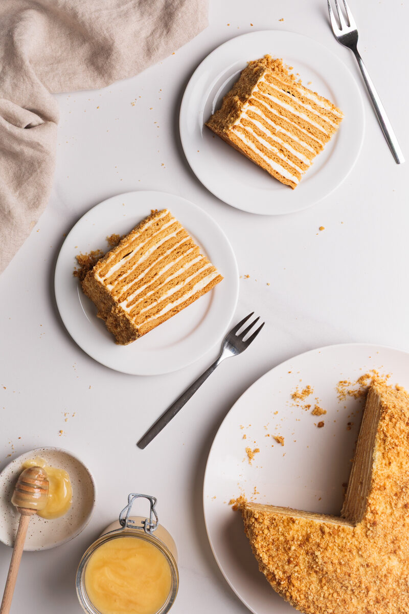 Top-down view of two plates with slices of layered honey cake, a whole cake with a missing slice, a small bowl of honey with a dipper, and a beige napkin on a white surface.