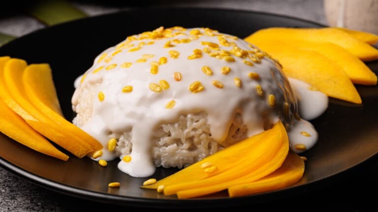 A serving of Thai mango sticky rice topped with coconut cream and sprinkled with yellow mung beans, garnished with sliced ripe mango on a black plate.