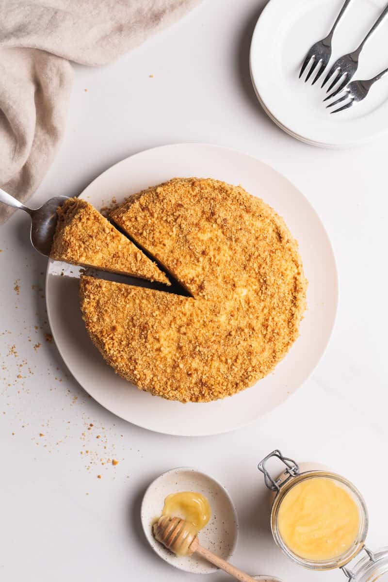 A top-down view of a round medovik cake on a white plate, with a slice being lifted. Nearby are stacked plates with forks, a jar of lemon curd, a small dish of honey, and a beige cloth.